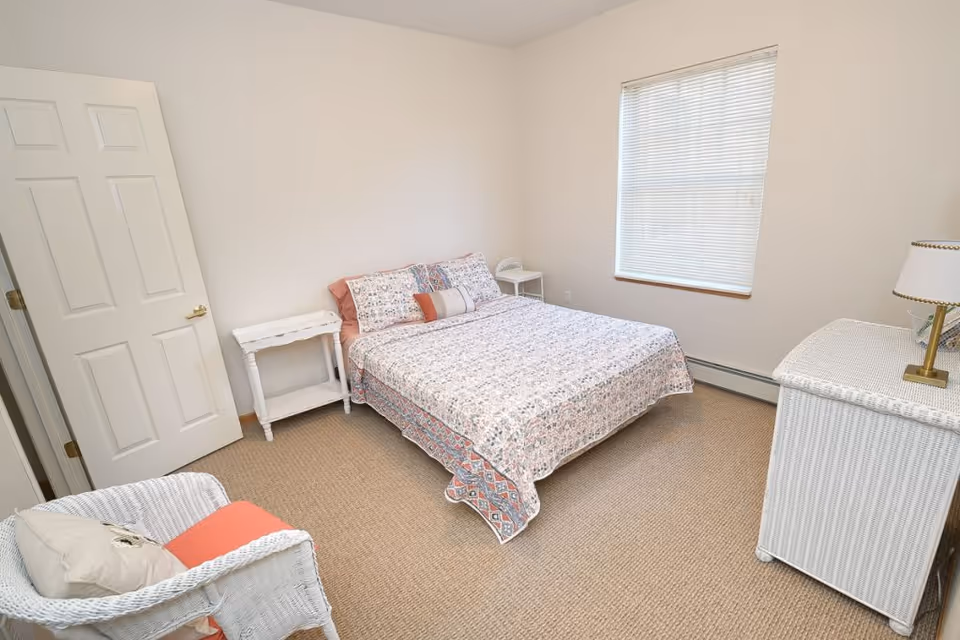 Simple bedroom with a double bed dressed in patterned bedding, white wicker furniture, a window with blinds, and a chair.