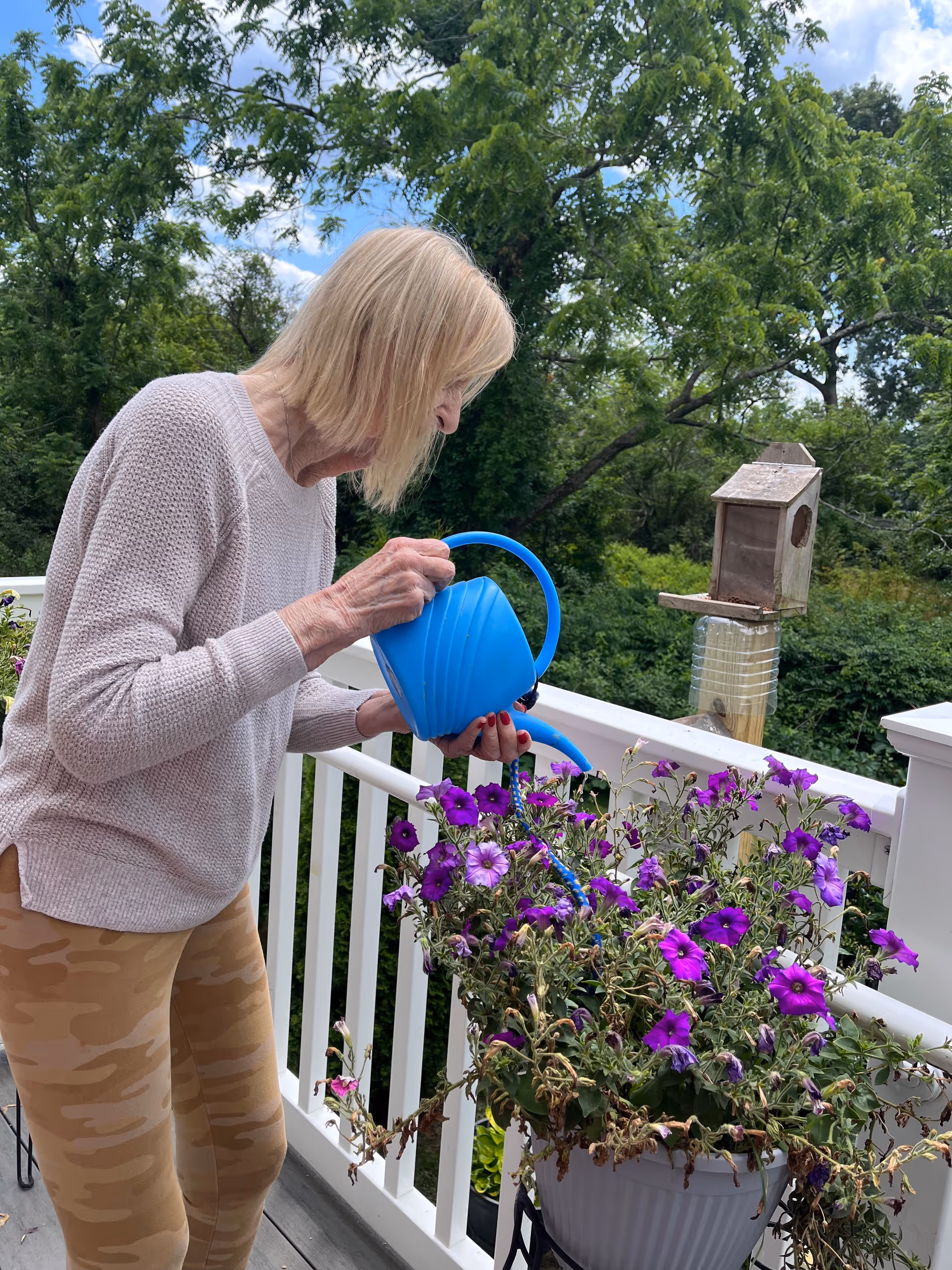 An elderly woman with blonde hair wearing a light pink sweater and beige camouflage leggings is watering purple flowers in a white planter on a balcony. The balcony has white railings, and there is a wooden birdhouse mounted on a post in the background with green trees and a partly cloudy sky.