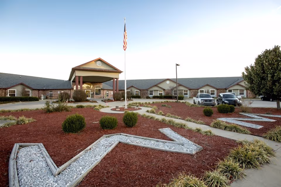 Exterior view of Emerald Care Center Claremore, showing a single-story building with a covered entrance, an American flag on a flagpole in front, landscaped garden beds with mulch and small bushes, and a parking area with several vehicles.