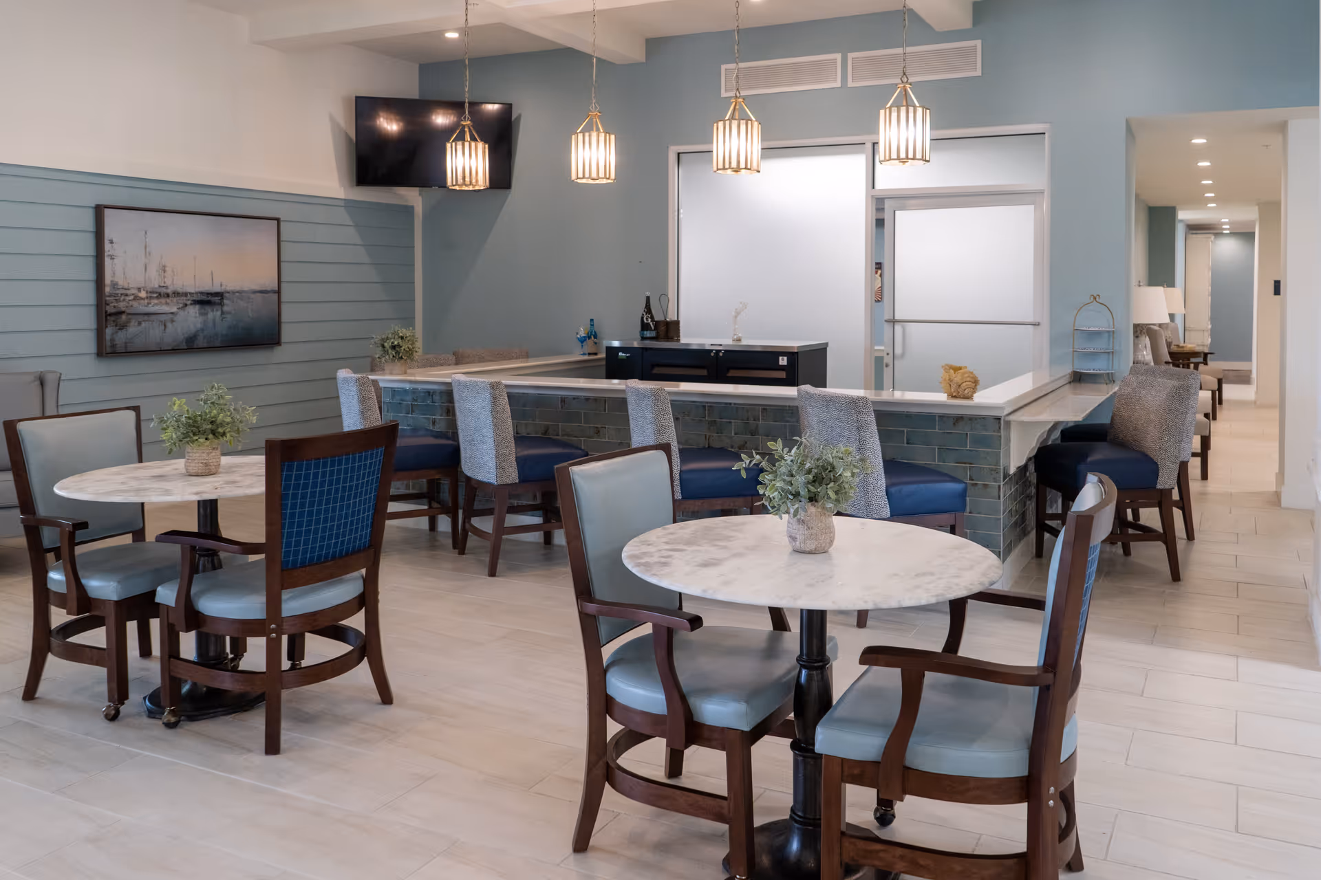 A modern dining area with round marble tables and wooden chairs with blue cushions. There is a bar counter with high chairs upholstered in blue and gray fabric. The walls are painted light blue with a painting of boats on one side and a flat-screen TV mounted above. Pendant lights hang from the ceiling, and there are plants on the tables adding a touch of greenery.