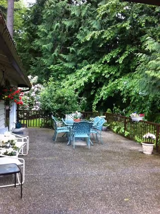 Outdoor patio area with a round glass table and six blue chairs surrounded by lush green trees and plants. There are flower pots with colorful flowers on the wooden railing and near the house wall. The patio surface is made of concrete, and part of the house with white siding and a hanging flower basket is visible on the left side.