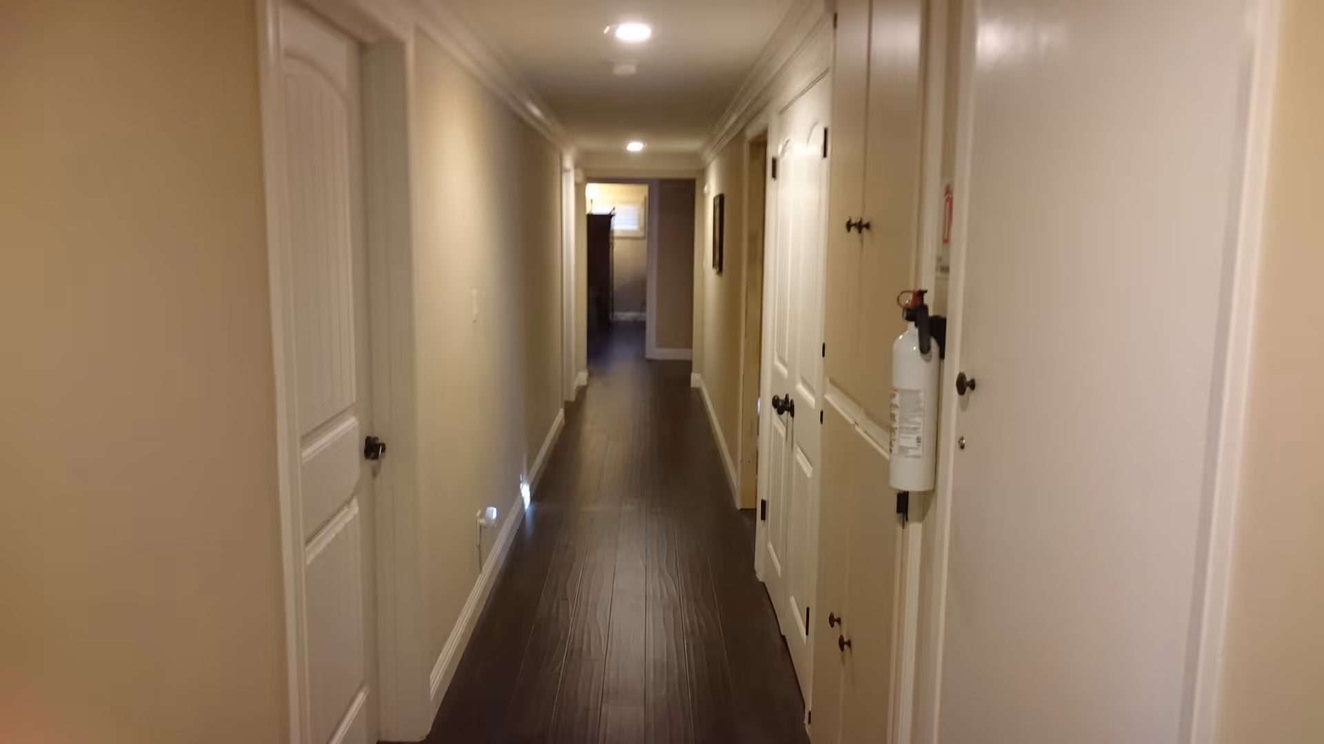 A long, narrow hallway with beige walls and dark wooden flooring. Several white doors line both sides of the hallway, and a fire extinguisher is mounted on the right wall. Recessed ceiling lights illuminate the corridor, and a small window is visible at the far end.