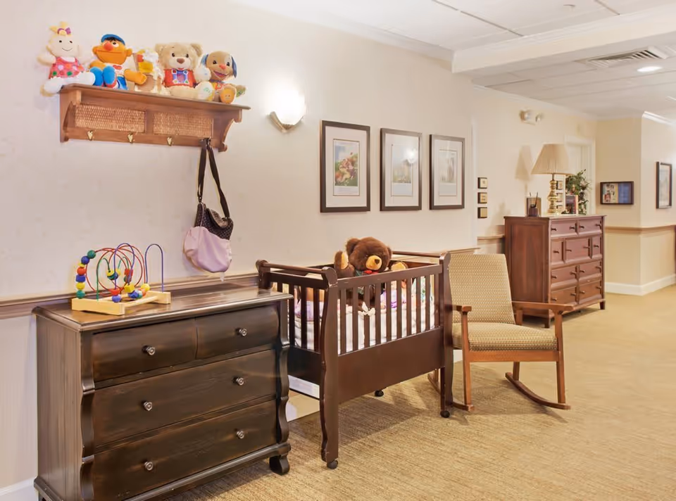 A cozy interior hallway area featuring a dark wooden dresser with a colorful bead maze toy on top, a wooden crib with a large teddy bear inside, and a cushioned rocking chair. Above the dresser is a wooden wall shelf holding several stuffed animals and a hanging purse. The walls are decorated with framed pictures and soft wall sconces provide lighting.
