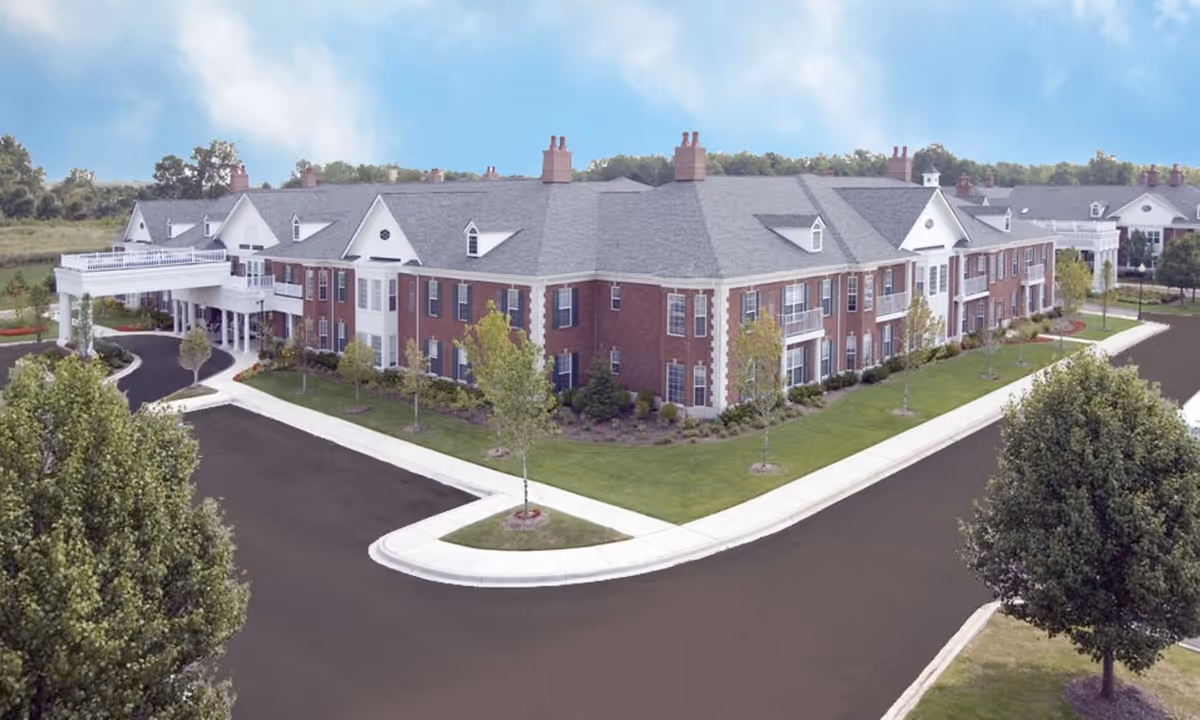 Exterior view of a large two-story senior living facility building with red brick walls, multiple windows, and white trim. The building has a covered entrance with white columns and a driveway. There are small trees and landscaped grass areas surrounding the building under a partly cloudy sky.