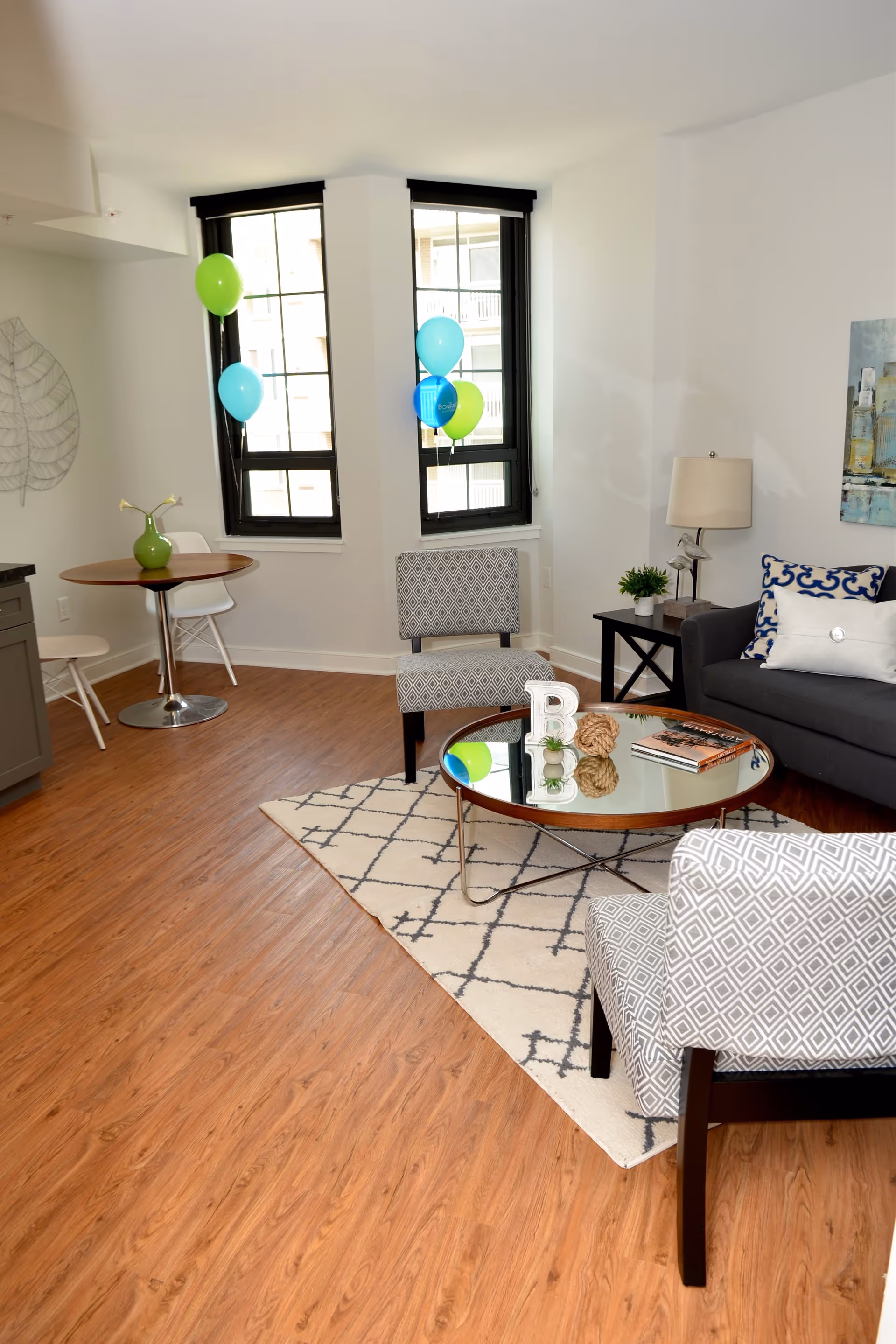 A bright living room with wooden flooring, two patterned armchairs, a gray sofa with decorative pillows, a round glass coffee table on a white rug with a geometric pattern, and a small round dining table with two white chairs near two tall windows with black frames. Blue and green balloons are tied near the windows, and there is a table lamp and a small plant on a side table next to the sofa.