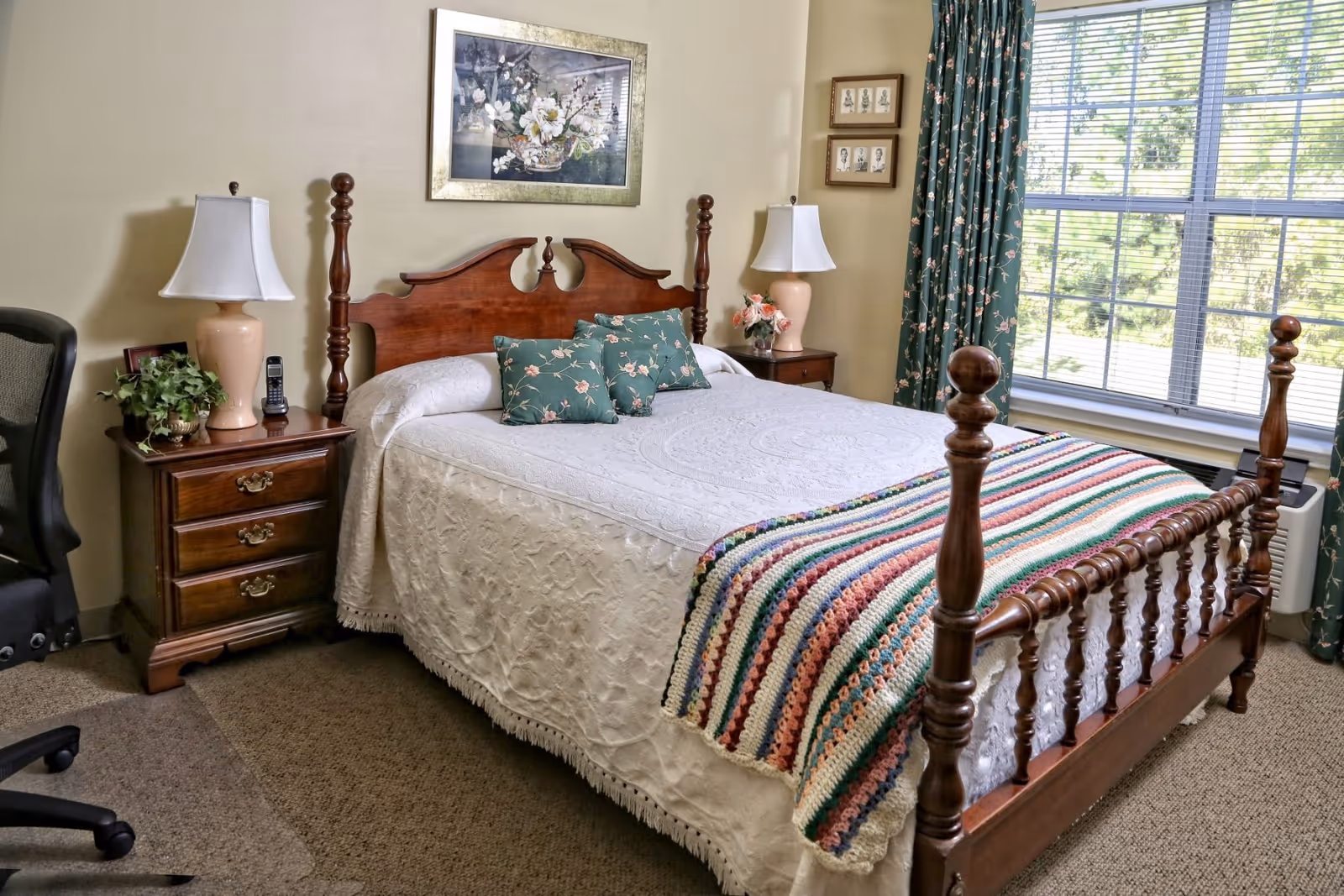 A cozy bedroom in Woodleaf Senior Care featuring a wooden bed with a white bedspread and a colorful crocheted blanket at the foot. There are three floral-patterned pillows on the bed. On each side of the bed are wooden nightstands with matching lamps, a plant, and a phone. The room has a large window with floral curtains letting in natural light, and framed artwork on the walls.