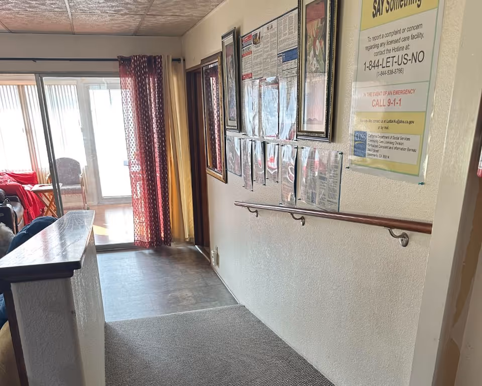 Interior hallway of a senior living facility with a wall-mounted handrail and posted notices, leading to sliding glass doors opening to a sunroom with seating.
