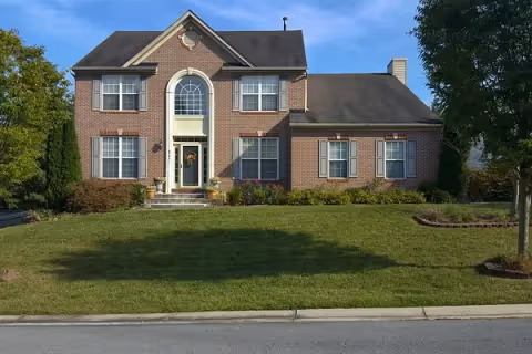 Front exterior view of a two-story brick house with a large arched window above the front door, multiple windows with white shutters, a well-maintained lawn, and some bushes and trees around the property under a clear blue sky.
