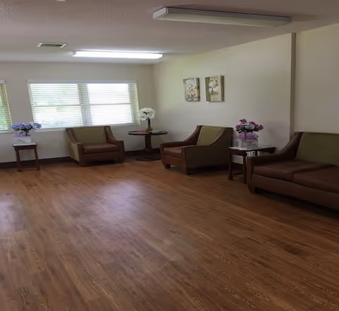A spacious room with wooden flooring and beige walls featuring three brown armchairs and a brown sofa with green cushions. There are three small tables with flower arrangements placed near the seating. Two floral paintings hang on the wall above one armchair. A large window with horizontal blinds allows natural light into the room.