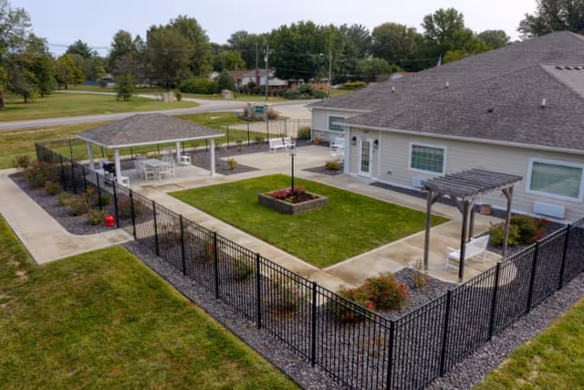 Outdoor fenced courtyard area with a central grassy lawn, a flower bed, a covered seating area with tables and chairs, and a pergola with a bench. The courtyard is adjacent to a single-story building with light-colored siding and several windows. Trees and a road are visible in the background.