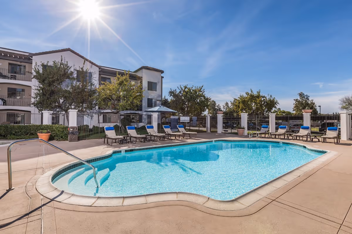 Outdoor swimming pool with clear blue water surrounded by a concrete deck with lounge chairs that have blue cushions. Behind the pool is a three-story building with balconies and trees, under a bright sunny sky.