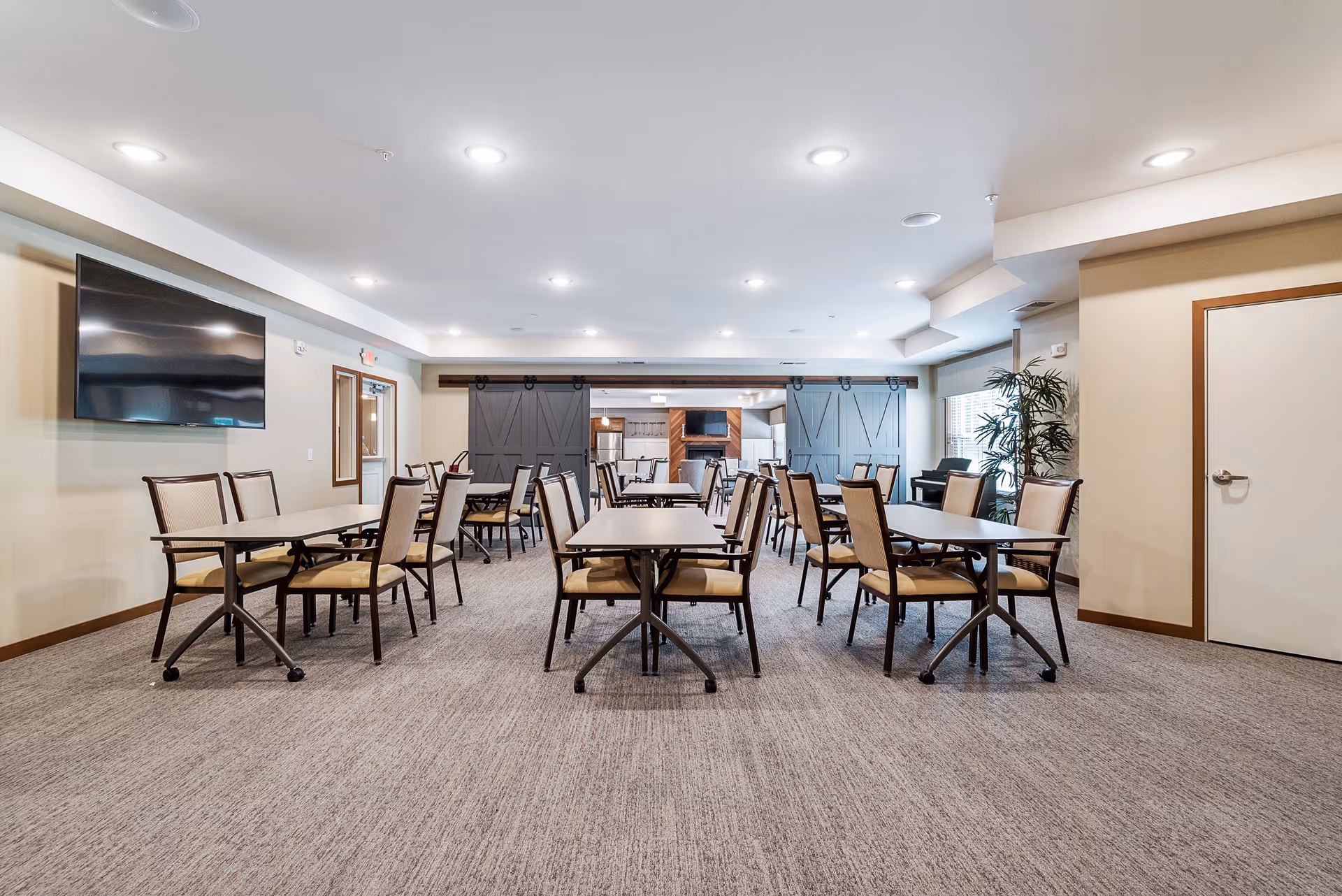 Spacious senior dining room with multiple tables and chairs, a wall-mounted TV, and sliding barn doors at the far wall.