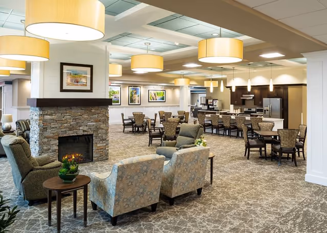 A spacious and well-lit common area in a senior living facility featuring a stone fireplace, comfortable armchairs, multiple dining tables with chairs, and a kitchen area with modern appliances in the background. The room has large hanging light fixtures and framed artwork on the walls.
