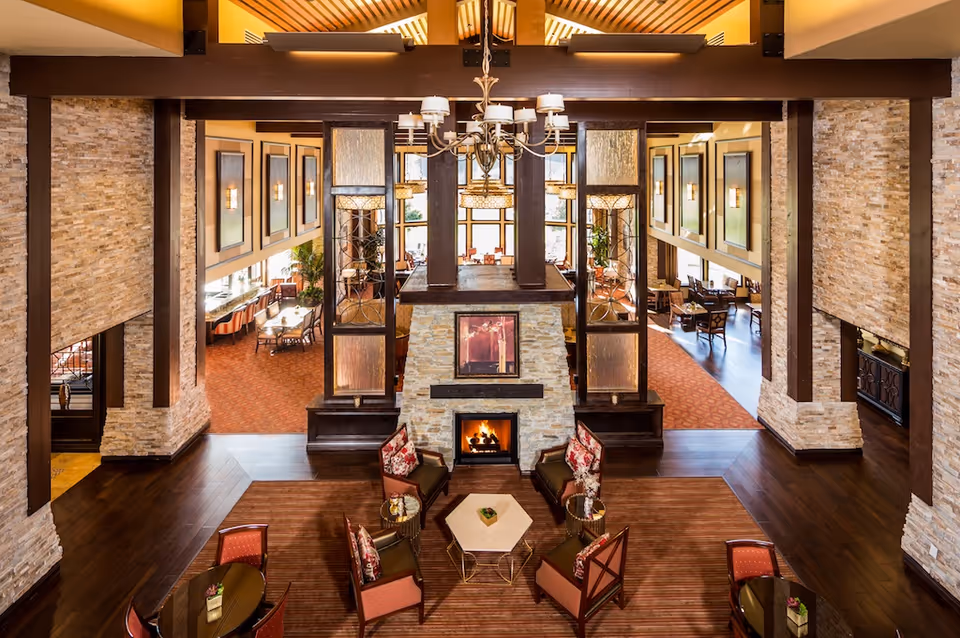 Spacious and warmly lit common area in a senior living facility featuring a central stone fireplace with a painting above it, surrounded by comfortable armchairs with floral cushions and small tables. The room has high ceilings with exposed wooden beams, large windows letting in natural light, and adjacent dining areas with tables and chairs visible in the background.