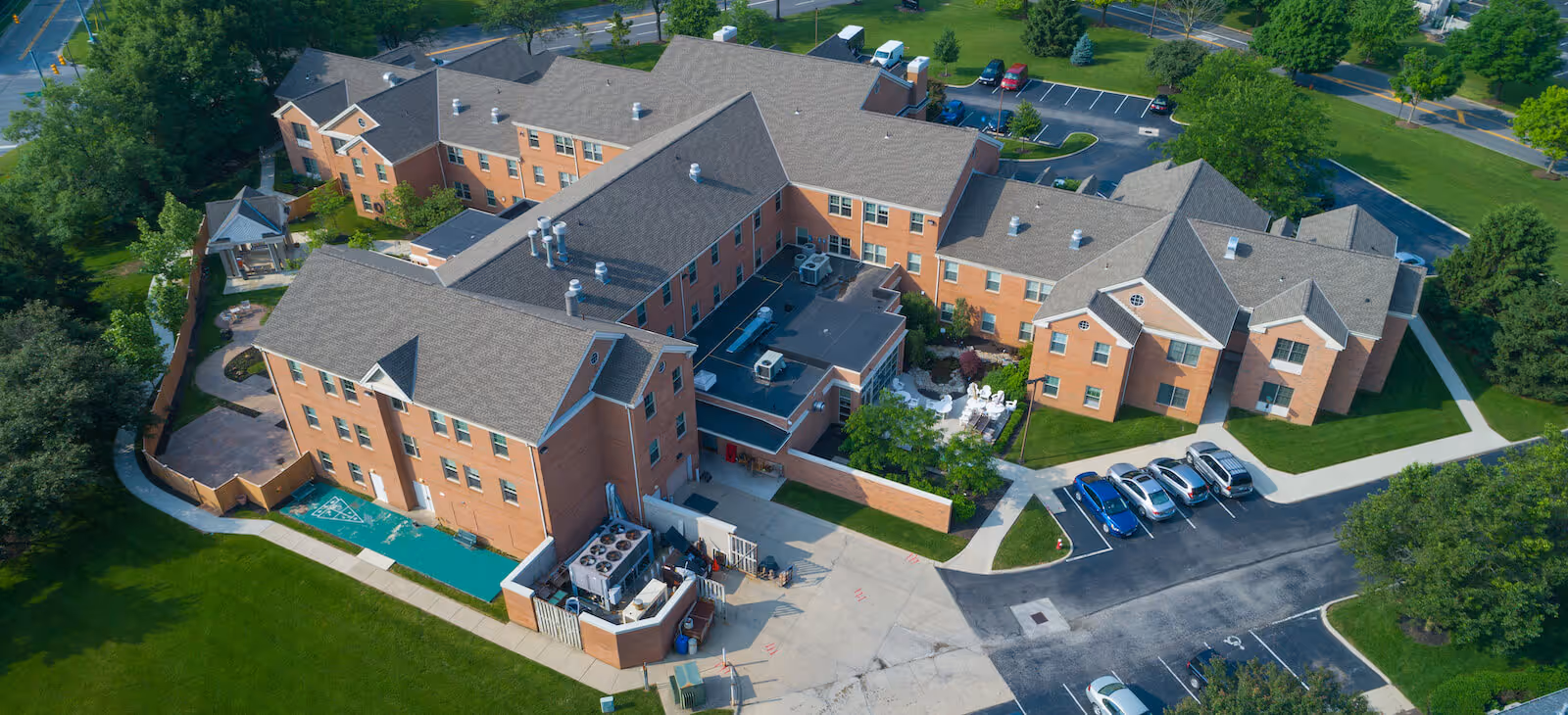 Aerial view of a large senior living facility named Harmony Trace, showing multiple connected brick buildings with gray roofs, surrounding green lawns, parking areas with cars, and paved walkways.