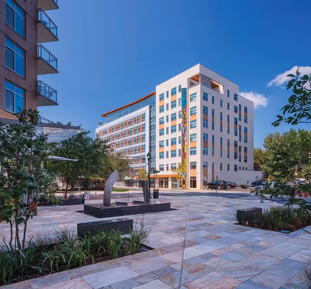 Modern multi-story residential building facing a tiled plaza with a small fountain sculpture, landscaping, and a clear blue sky.