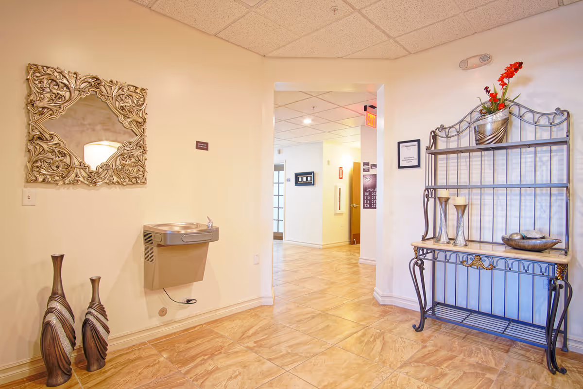 Interior hallway of an assisted living facility featuring a decorative mirror, a wall-mounted drinking fountain, floor vases, and a metal display shelf with decor.