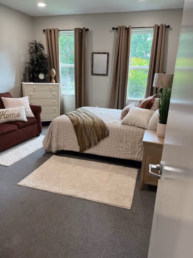 A cozy bedroom with a neatly made bed covered in a white quilt and a brown patterned throw blanket. There are several pillows on the bed. To the right of the bed is a wooden nightstand with a lamp and a potted plant. On the left side of the room, there is a maroon armchair with a white pillow that says 'Home' and a white dresser with decorative items including a clock and a plant. Two windows with beige curtains let in natural light, and a small framed mirror hangs on the wall between the windows. The floor is carpeted with two white rugs.