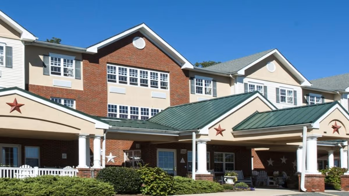 Front exterior view of a multi-story senior living facility building with brick and beige siding, green metal roofs over the entrance areas, white columns, decorative stars, and well-maintained shrubs and greenery in front under a clear blue sky.