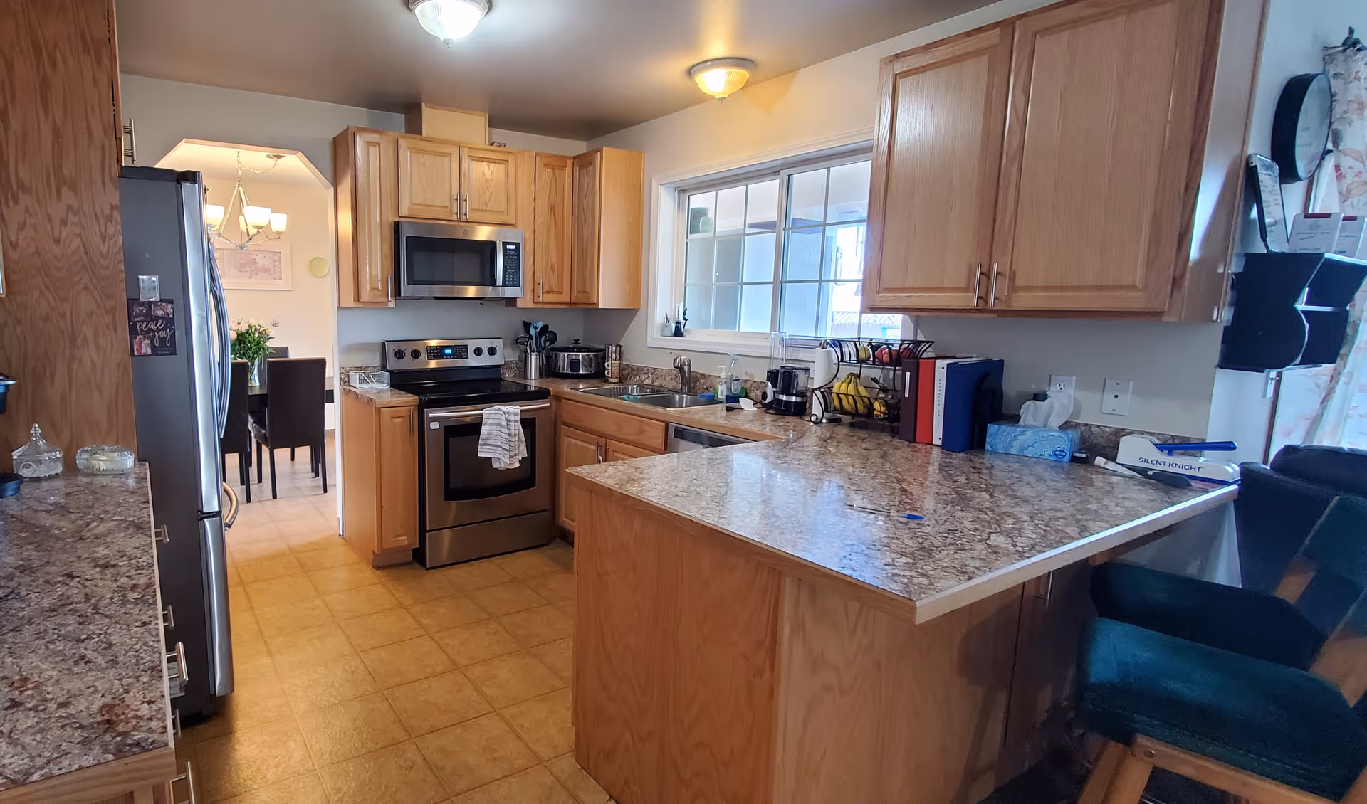 Bright kitchen with wooden cabinets, granite countertops, stainless steel stove and microwave, and a breakfast bar with chairs.