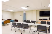 A room with multiple chairs arranged around tables facing a wall-mounted television. In the background, there are hospital-style beds and a privacy curtain, suggesting a medical or care facility setting.