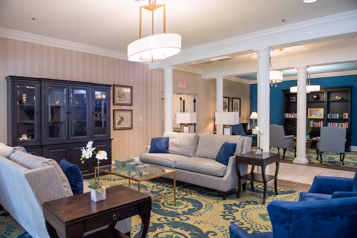 A spacious and elegantly decorated senior living common area featuring light gray sofas with blue accent pillows, dark wood side tables with white orchids, a glass coffee table, and a large patterned area rug. The room includes white columns, a dark wood cabinet with glass doors, framed artwork on the walls, and a separate seating area with two patterned armchairs and a bookshelf filled with books against a blue accent wall.