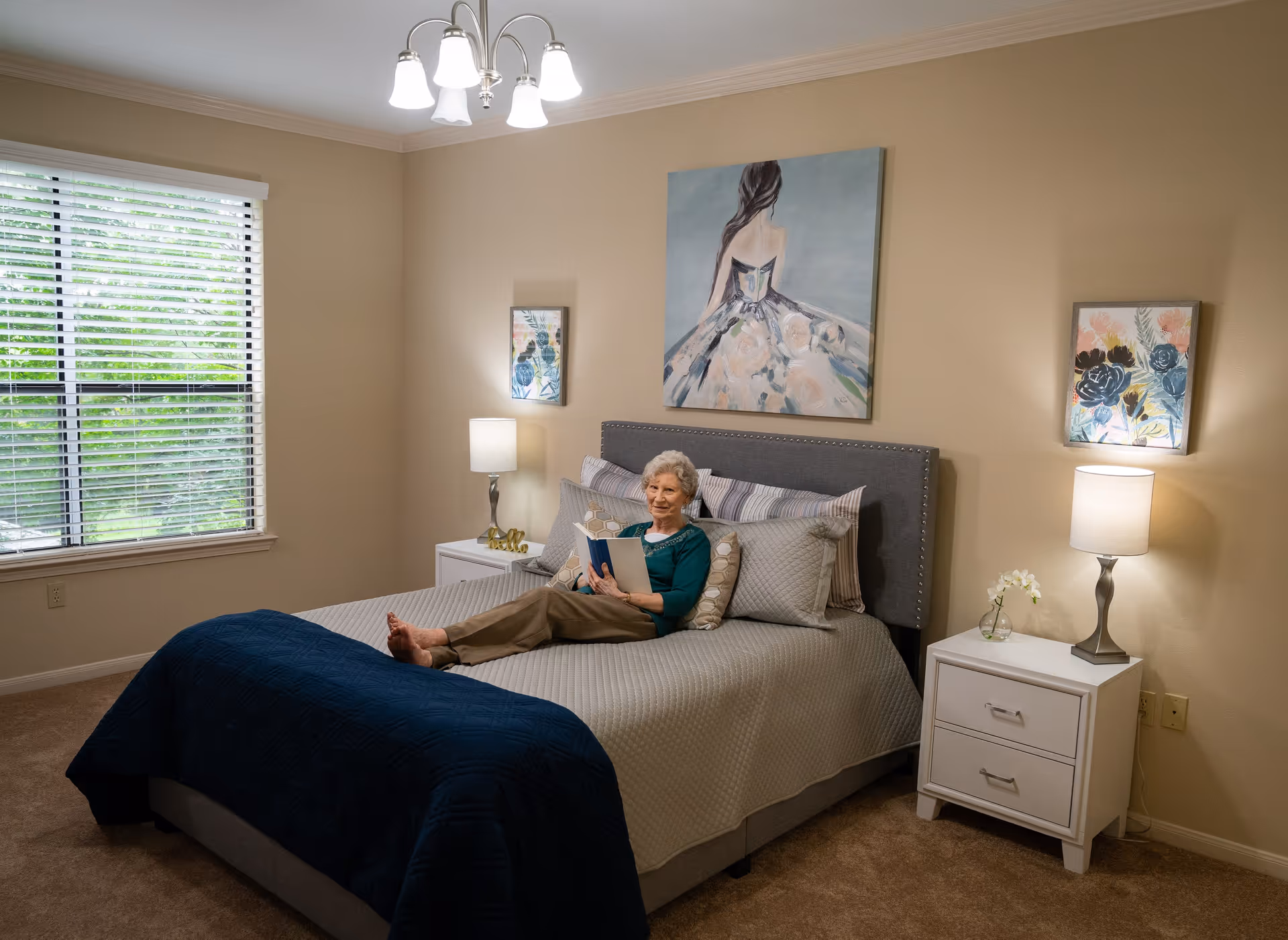 An elderly woman is lying on a neatly made bed in a cozy bedroom. The room features beige walls, a large window with white blinds, two white nightstands with lamps, and three pieces of artwork on the walls. The bed has a gray quilted bedspread with a dark blue blanket folded at the foot.