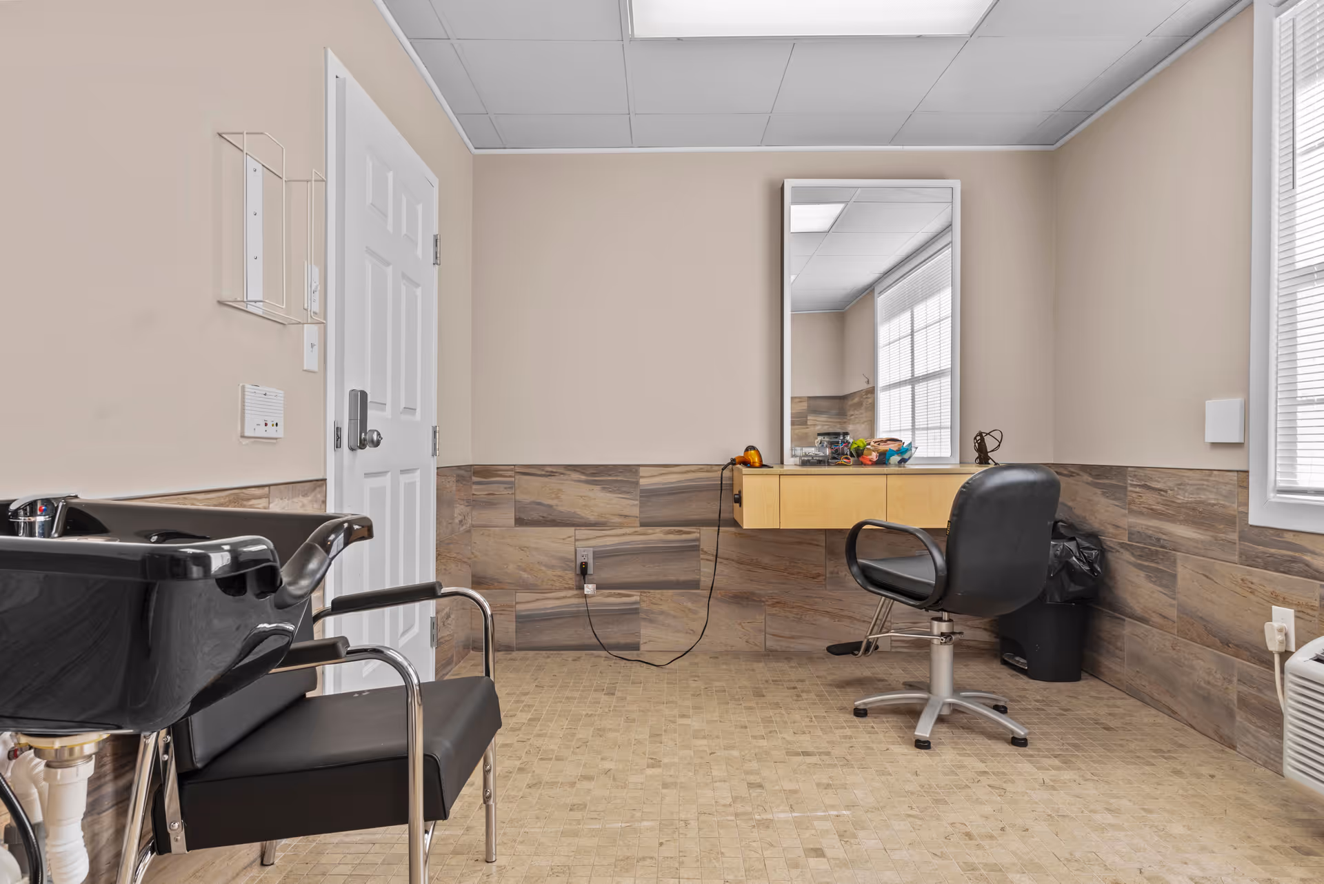 A small salon room with a black shampoo chair and sink on the left, a black salon chair in front of a large mirror mounted on the wall, a floating wooden shelf beneath the mirror with various hair styling tools, beige tiled floor and walls with a beige painted upper wall, and a window with blinds on the right.