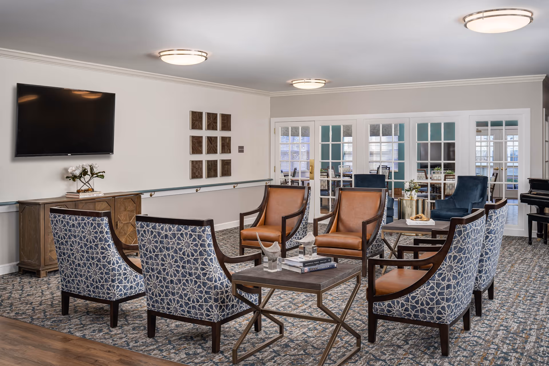 A senior living facility common area with a seating arrangement consisting of six chairs around a coffee table. Four chairs have a blue and white patterned upholstery, and two chairs have brown leather upholstery. The room features a carpeted floor, a wall-mounted flat-screen TV, a wooden cabinet with a small plant, and decorative wall art. In the background, there are glass-paneled doors leading to another room with additional seating and a piano on the right side.