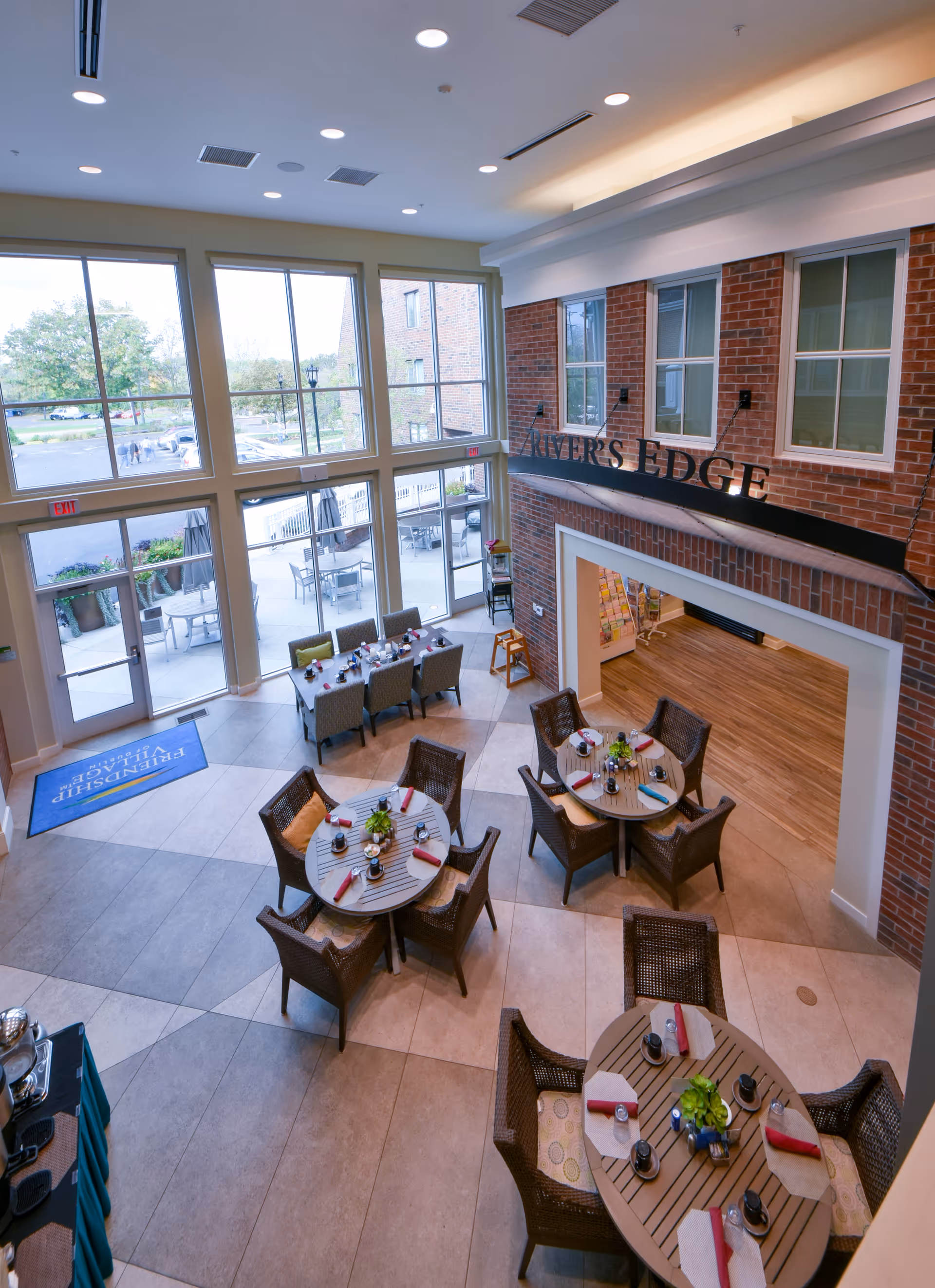 Interior view of a dining area in a senior living facility with round and rectangular tables set with plates, napkins, and condiments. The space features large floor-to-ceiling windows letting in natural light, a brick wall with the sign 'RIVER'S EDGE,' and an open doorway leading to another room. Outside, there is a patio area with tables and umbrellas.