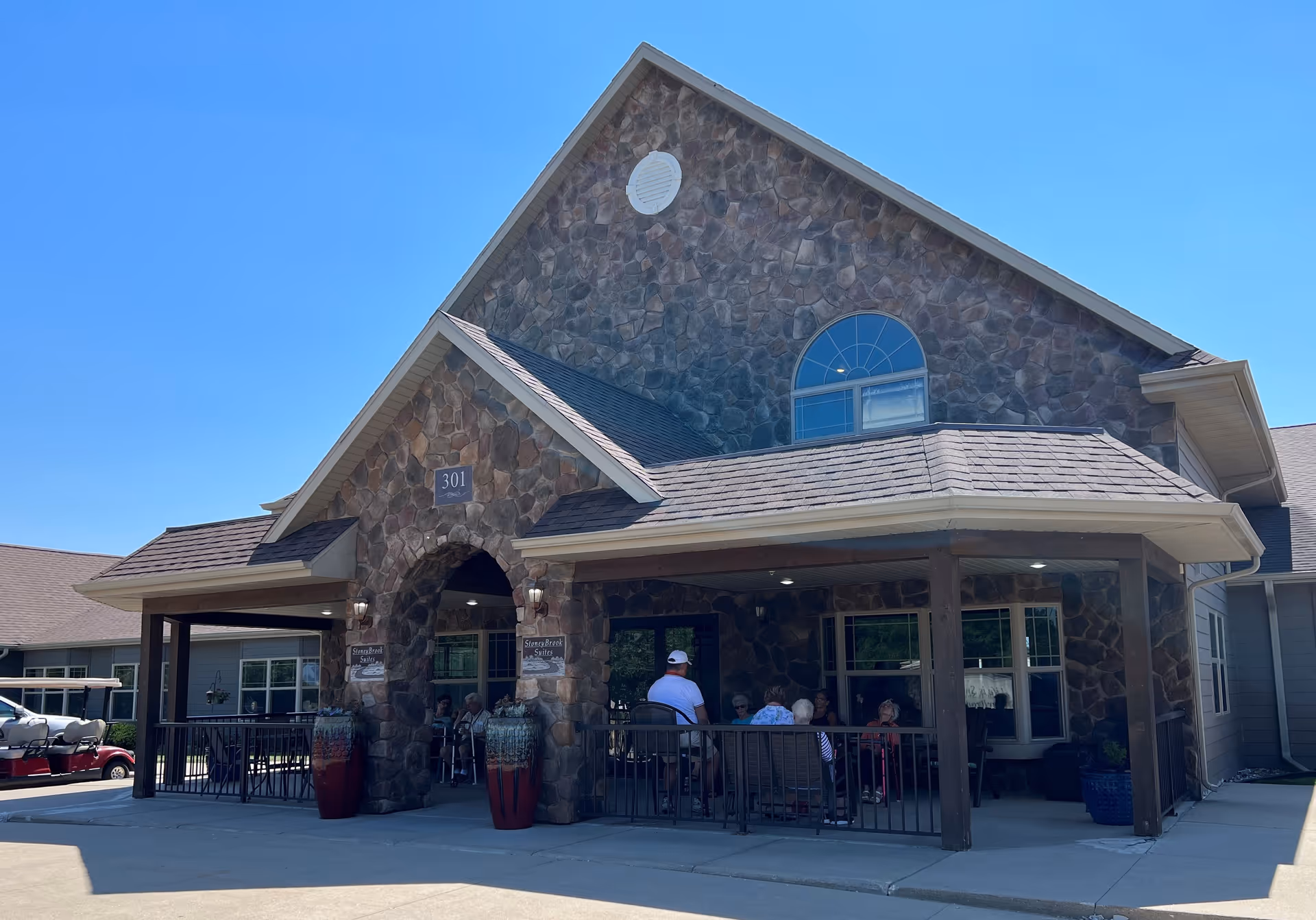 Exterior view of StoneyBrook Suites Assisted Living building with stone facade and covered porch. Several people are seated on the porch under the roof, enjoying the outdoor seating area. The sky is clear and blue.