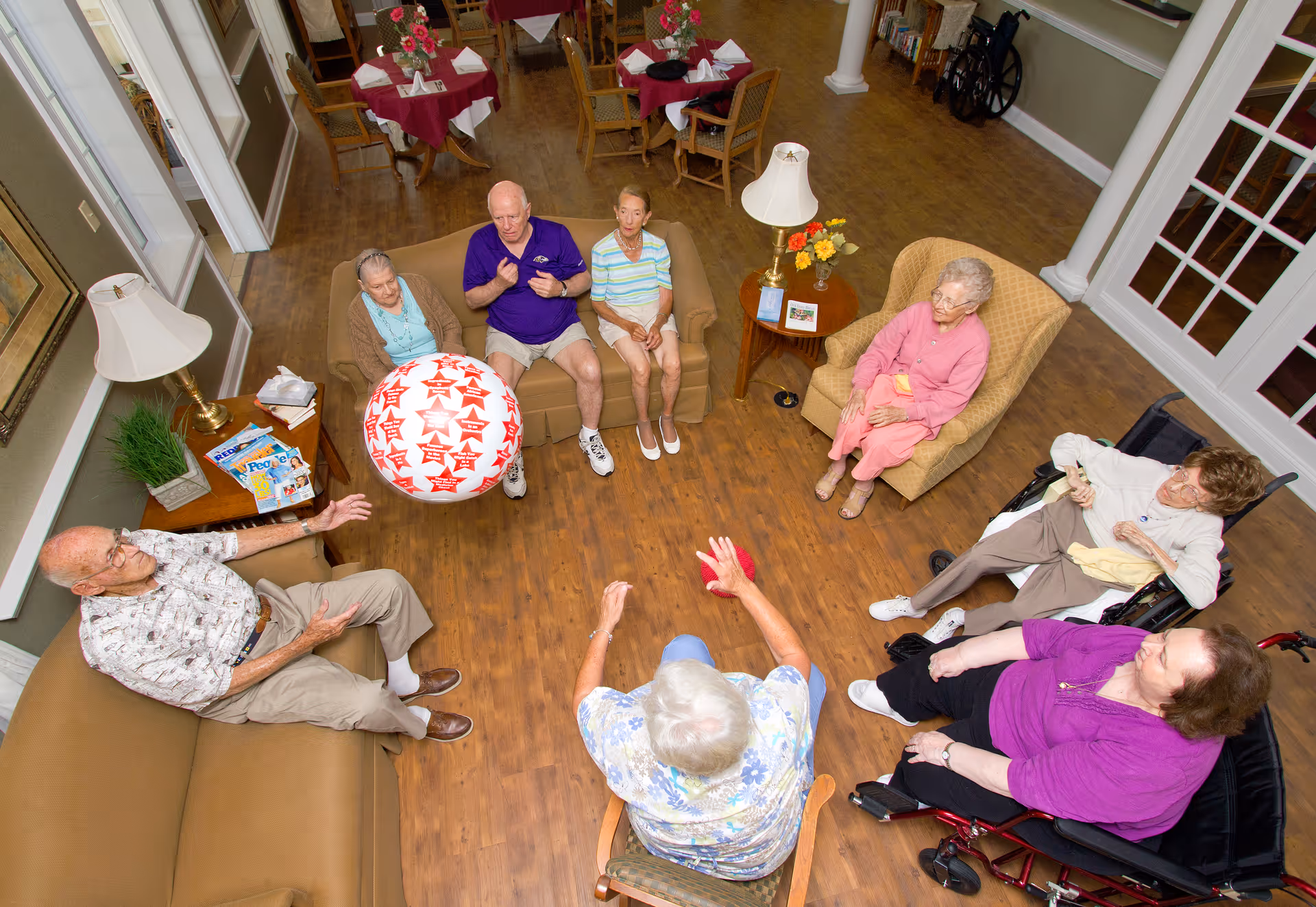A group of elderly people sitting in a circle in a living room area, engaging in a seated activity with a large inflatable ball. The room has wooden flooring, comfortable chairs and sofas, side tables with lamps and flowers, and a wheelchair visible in the background.