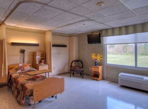 Hospital-style single bed in a senior living room with dresser, chair, flowers, and window.