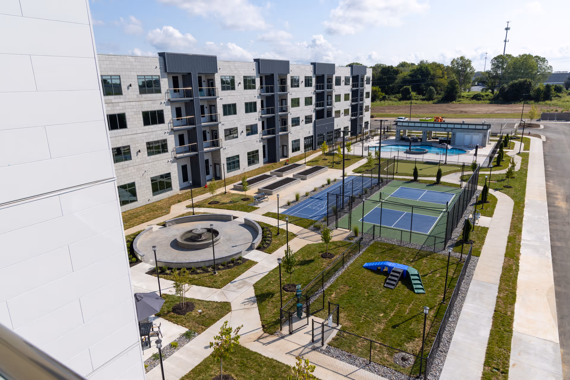 View of an outdoor recreational area at The Preston Luxury Living featuring a circular water fountain, fenced tennis and pickleball courts, a swimming pool with a covered seating area, and a small playground on a grassy lawn, surrounded by a modern multi-story residential building.