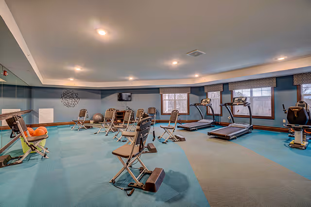 A fitness room with several exercise chairs, treadmills, an exercise bike, and a rack of orange exercise balls. The room has blue carpeting, large windows with valances, and a wall-mounted TV. There is a large mirror on one wall and ceiling lights illuminating the space.