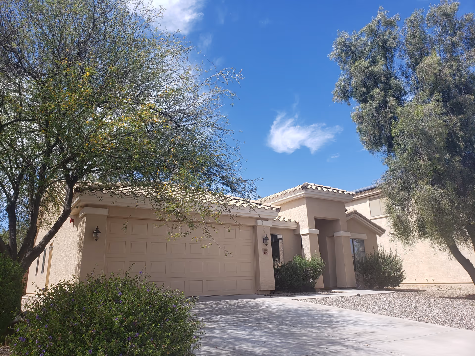 Exterior view of a single-story beige stucco house with a tiled roof, a two-car garage, and a driveway. There are trees and shrubs in the front yard under a clear blue sky with a few clouds.