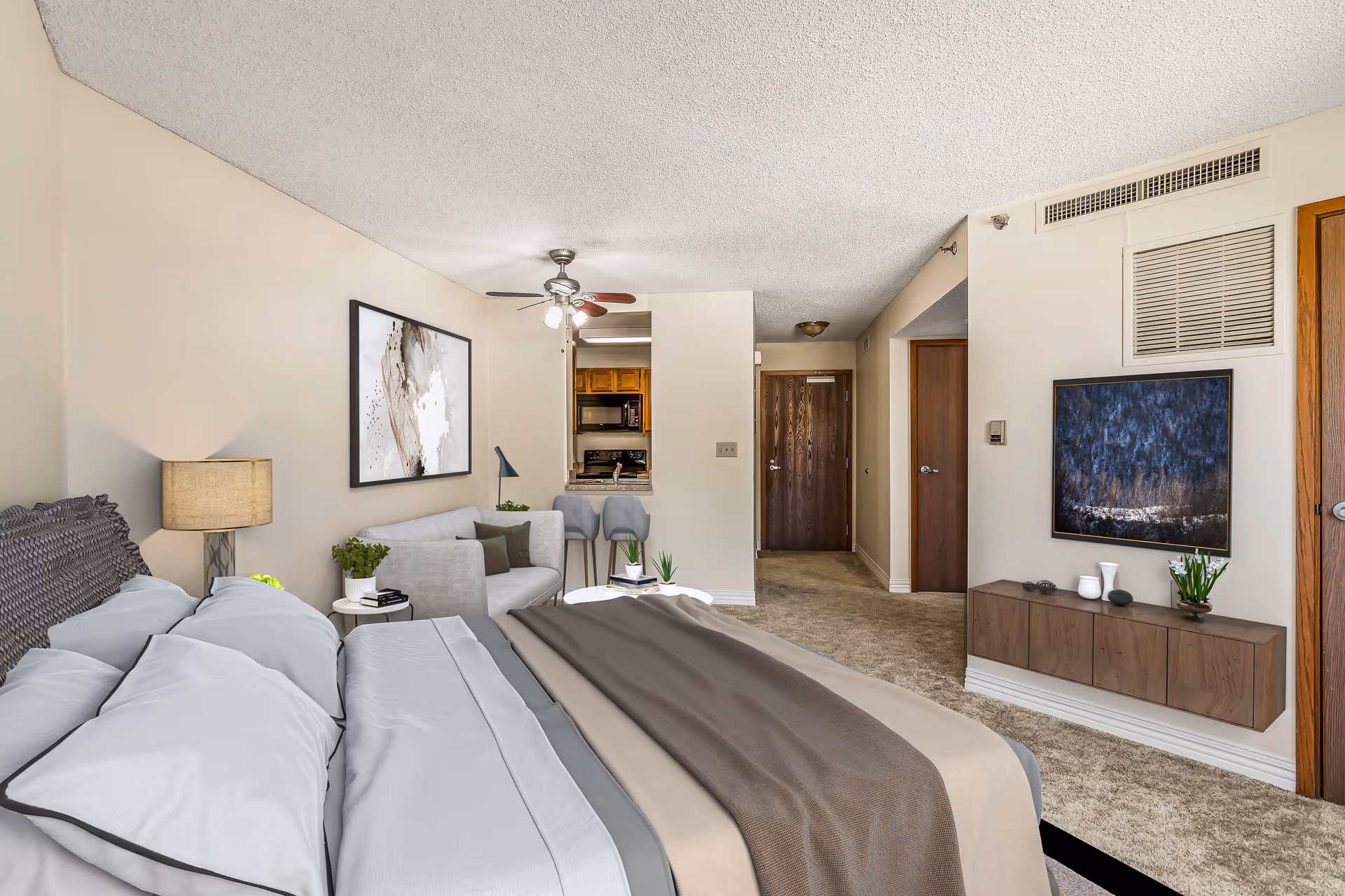 A well-lit bedroom with a large bed featuring white and gray bedding and a brown throw blanket. To the left of the bed is a nightstand with a lamp and a small plant. A gray sofa with cushions is positioned against the wall, beneath a large abstract painting. The room has beige walls and carpeted floors. In the background, there is a small kitchen area with two bar stools and wooden cabinets. On the right wall, a flat-screen TV is mounted above a floating wooden cabinet with decorative items and a small plant. Two wooden doors and an entrance door are visible in the hallway.