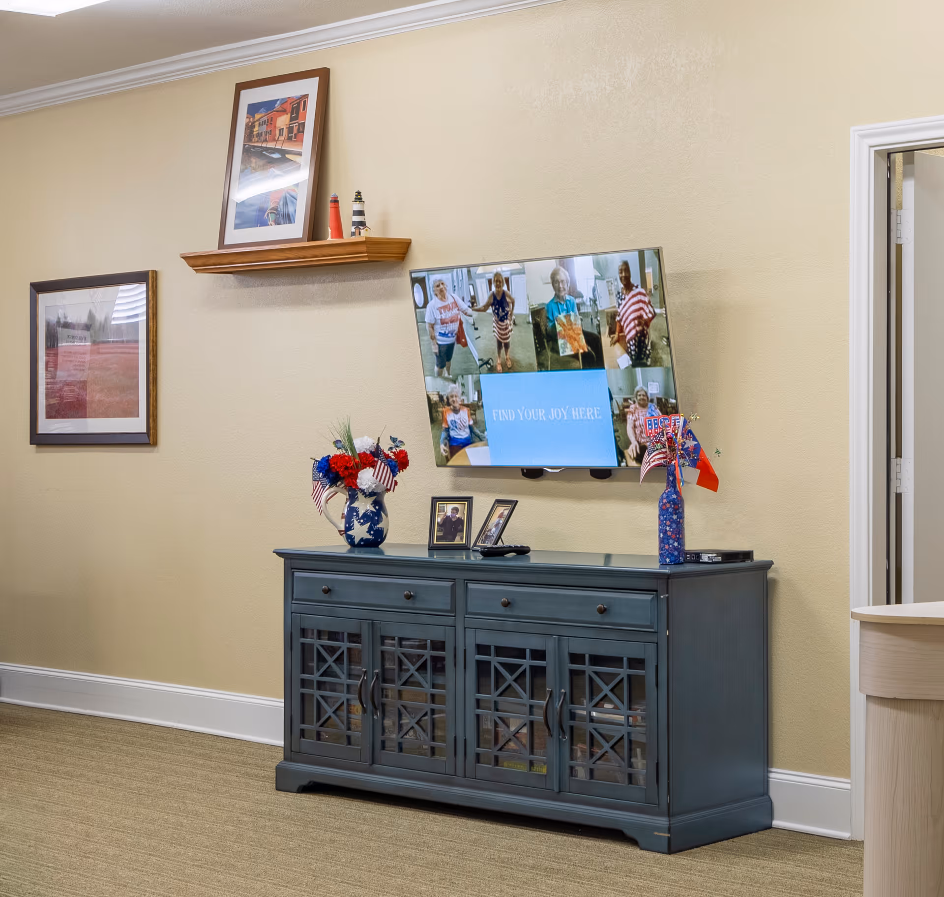 Interior view of a room with a blue cabinet against a beige wall. On top of the cabinet are two vases with red, white, and blue floral arrangements and small American flags, along with two framed photos and a TV remote. Above the cabinet, a flat-screen TV is mounted on the wall displaying a collage of elderly people and the text 'FIND YOUR JOY HERE'. There are two framed pictures on the wall, one above a wooden shelf holding two small lighthouse figurines. A doorway is visible on the right side.