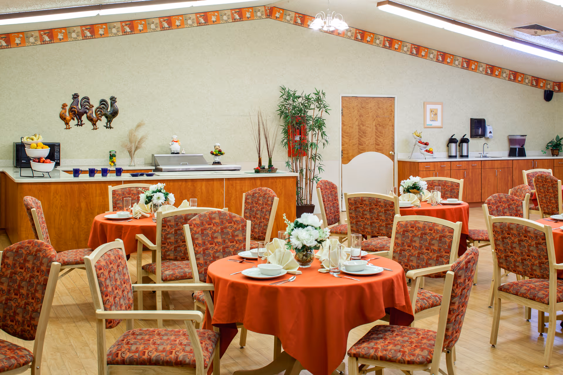 Dining room with round tables covered in orange tablecloths, each set with white plates, bowls, glasses, and napkins folded in a decorative manner. The chairs have wooden frames with patterned upholstery. The back wall features a counter with a microwave, fruit baskets, coffee dispensers, and decorative items including rooster wall art and plants. The room has a warm and inviting atmosphere.