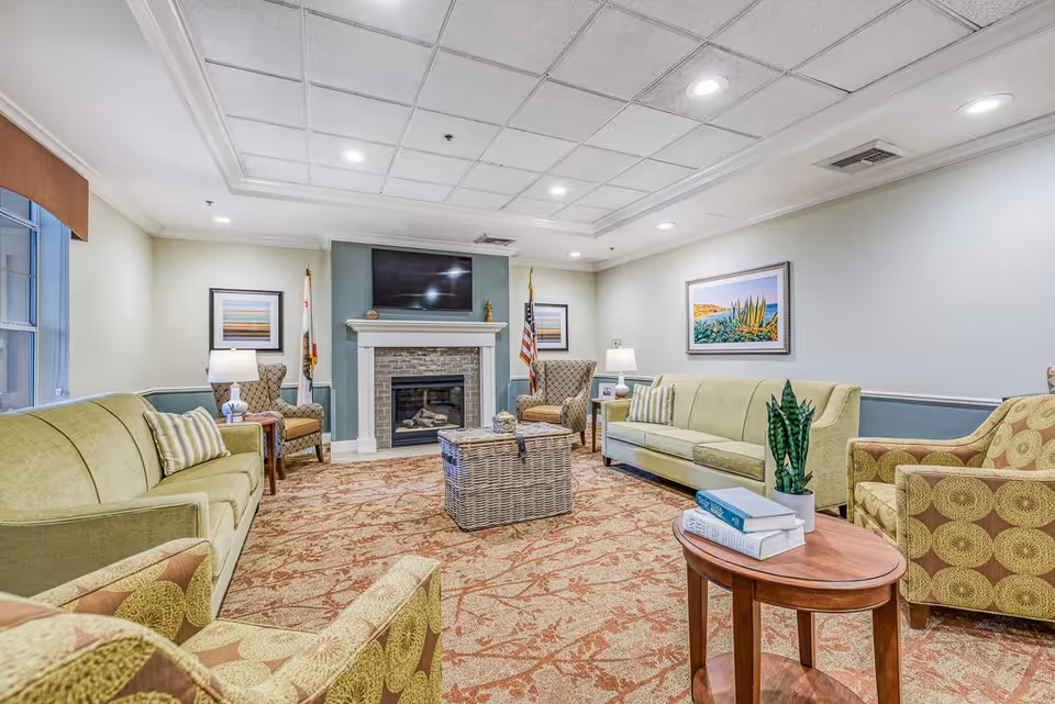 A cozy living room with two green sofas, two patterned armchairs, a wicker coffee table, and a wooden side table with books and a plant. The room features a fireplace with a TV mounted above it, two framed pictures on the walls, and a patterned carpet. The ceiling has recessed lighting.