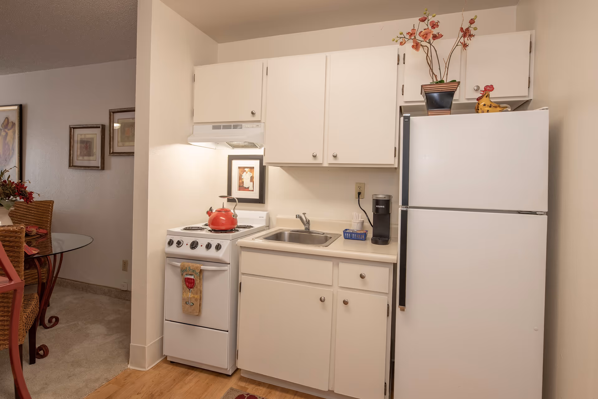 A small kitchen area with white cabinets, a white stove with a red kettle on top, a sink, a white refrigerator, and a Keurig coffee maker. There is a decorative plant and a ceramic chicken on top of the refrigerator. To the left, part of a dining area with a glass table and wicker chairs is visible.