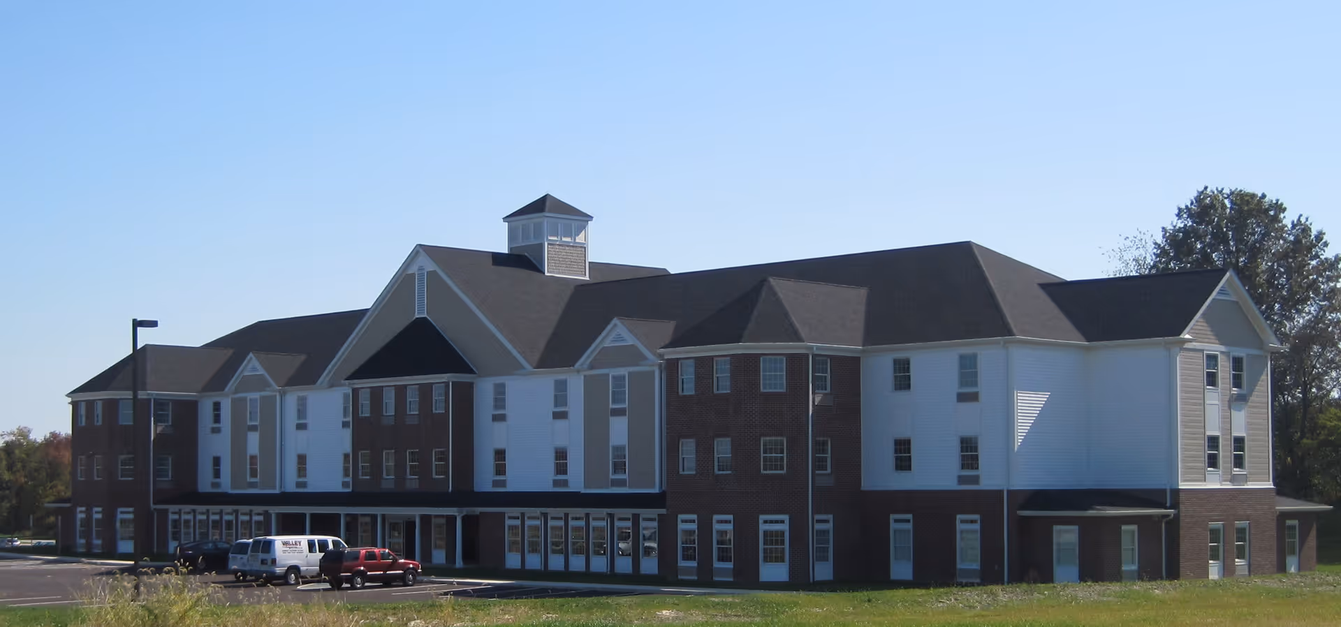 Front exterior of a three-story brick and white-siding assisted living building with a parking lot and grassy lawn.
