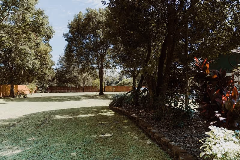 A spacious outdoor garden area with green grass, tall trees providing shade, and a flower bed with various plants along the right side. A wooden fence is visible in the background under a partly cloudy sky.