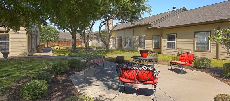 Outdoor patio area at Vista Ridge with metal chairs and benches featuring red cushions, surrounded by trimmed bushes and trees, adjacent to beige residential buildings under a clear blue sky.
