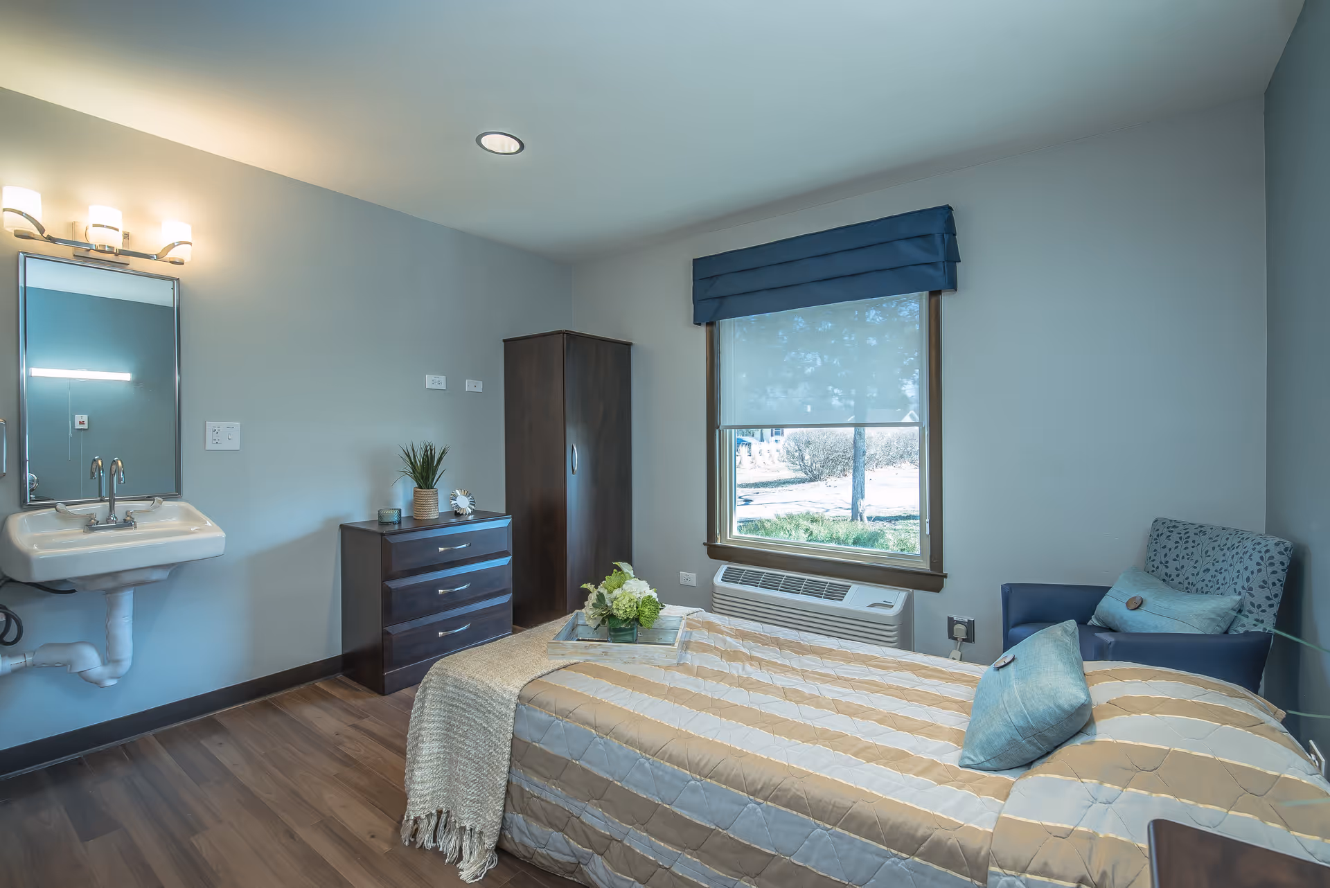 A neatly arranged bedroom in a senior living facility with a single bed covered in a beige and white striped quilt, a blue armchair with a pillow, a window with a blue valance and a roller shade, a dark wooden dresser and wardrobe, and a wall-mounted sink with a mirror and light fixture above it.