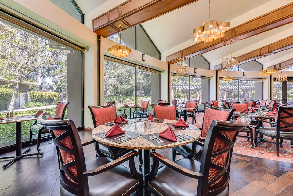 A bright dining room with large windows showing a garden outside. The room has round tables set with red cloth napkins, silverware, and glasses. The chairs have dark wood frames with red cushioned backs and seats. The ceiling is vaulted with wooden beams and multiple decorative chandeliers hanging down.