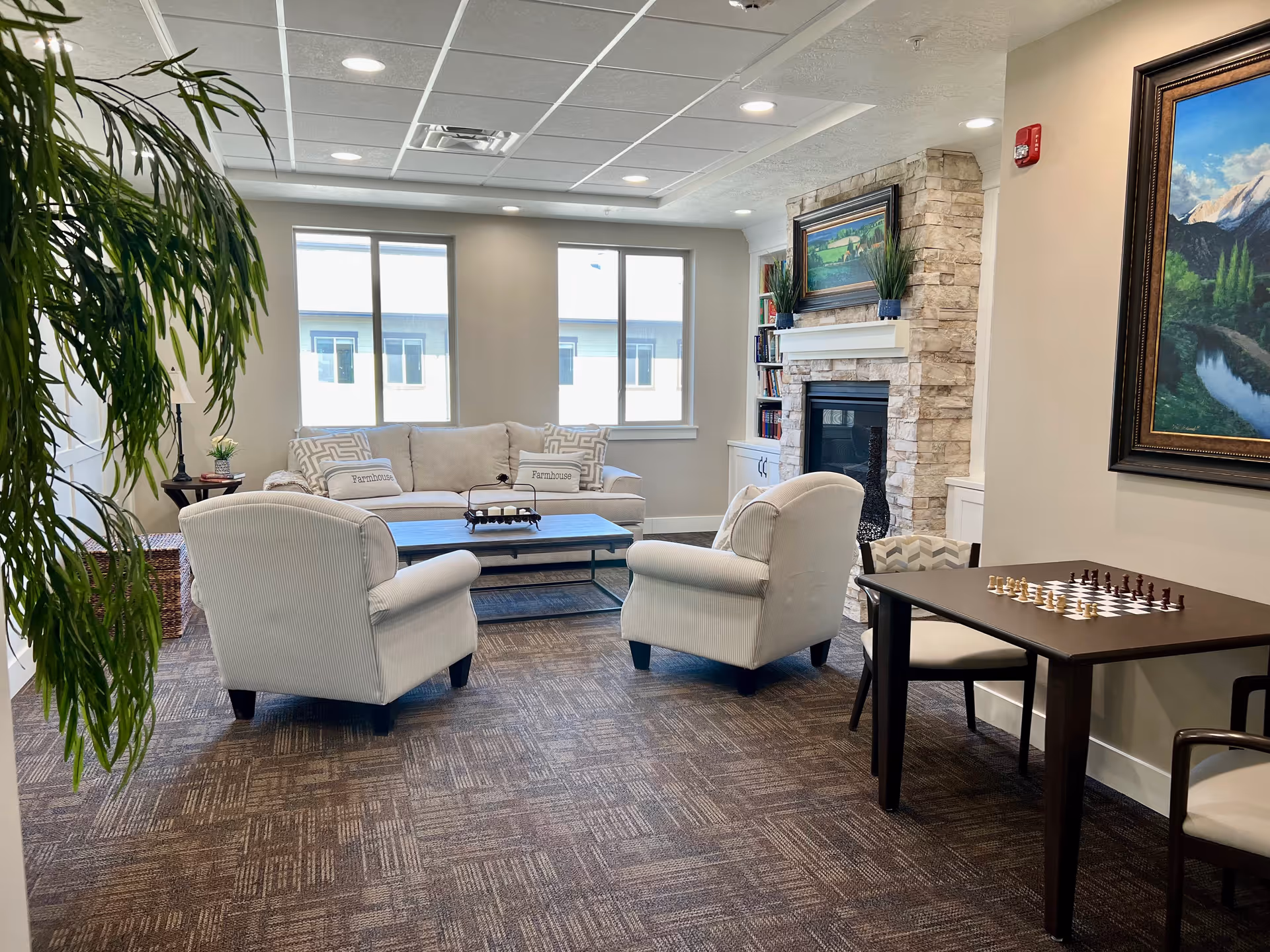 A cozy senior living common area with a beige sofa and two armchairs arranged around a coffee table. There is a stone fireplace with a painting above it and built-in shelves on either side. A small table with a chessboard and chairs is positioned near the wall, which also features a large framed landscape painting. The room has large windows letting in natural light and a potted plant in the corner.