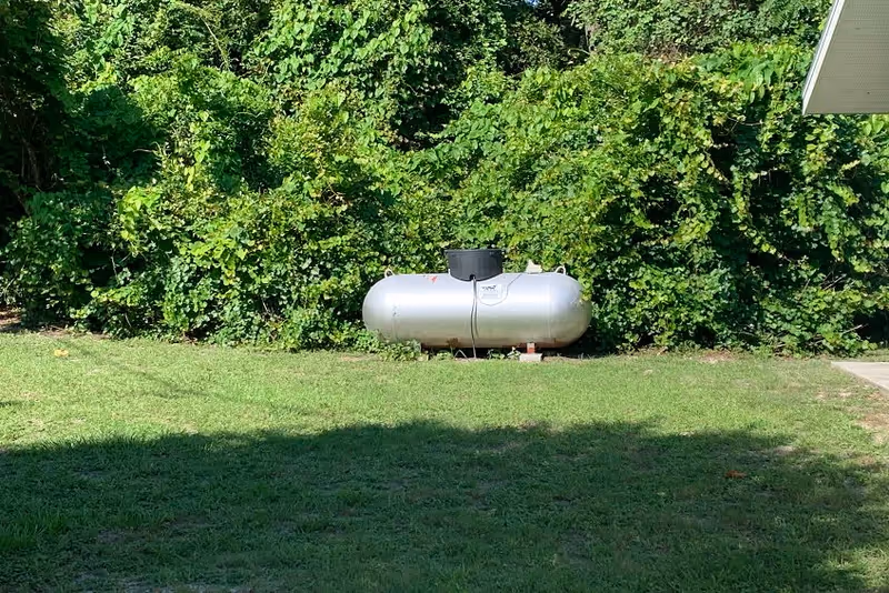 A large silver propane tank situated on a grassy lawn with dense green bushes and trees in the background. Part of a building's roof is visible on the right side of the image.