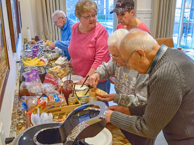 Several elderly residents serving themselves from a buffet table with breads, condiments and fruit in a communal dining area.