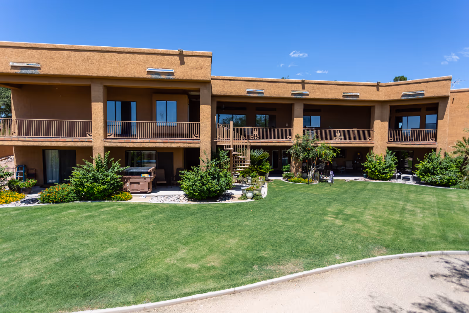 Exterior view of a two-story assisted living facility building with a brown stucco finish. The building has balconies on the upper floor and patios on the ground floor, surrounded by green bushes and a well-maintained lawn under a clear blue sky.